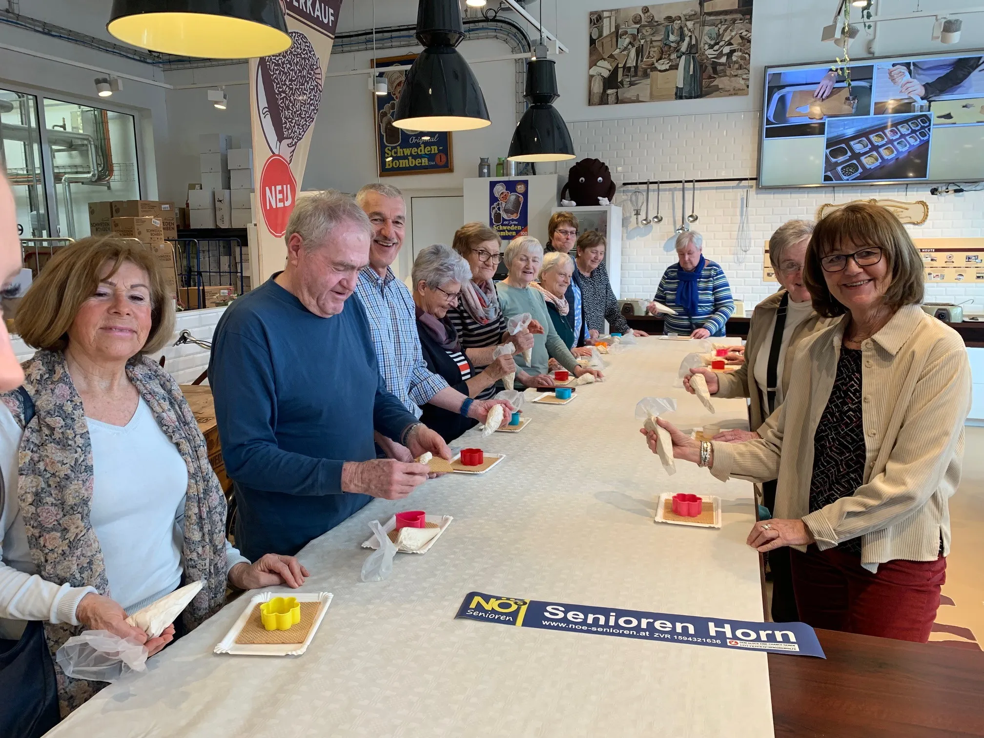 Eine Gruppe älterer Menschen steht um einen langen Tisch herum und bereitet Essen zu. Sie lächeln und scheinen sich zu amüsieren. Auf dem Tisch befinden sich Teller, Plastikverpackungen und ein Banner mit dem Text 'Senioren Horn'.