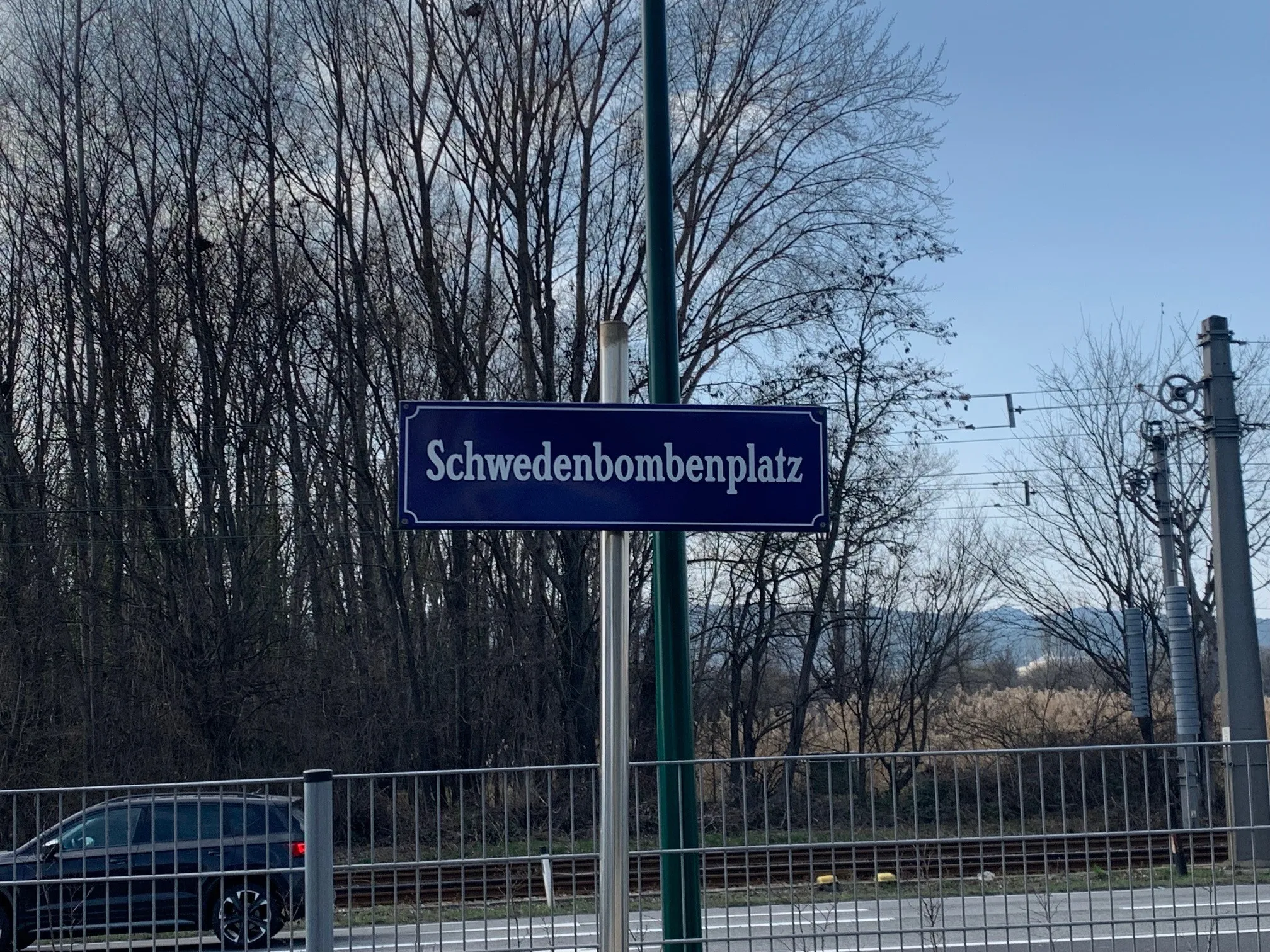 Ein blaues Schild mit der Aufschrift Schwedenbombenplatz ist an einem Pfosten montiert. Der Hintergrund zeigt Bäume und einen Zaun. Links ist ein Auto geparkt.