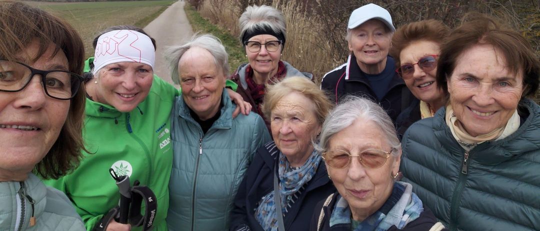 Eine Gruppe älterer Frauen mit Brillen, die auf einem ländlichen Pfad lächelnd für ein Foto posieren, mit Grünflächen und einem klaren Himmel im Hintergrund.