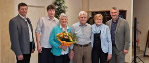 Eine Gruppe von Menschen steht zusammen in einem Raum. Eine ältere Frau hält einen Blumenstrauß. Der Mann und die Frau in der Mitte lächeln. Eine Kronleuchter hängt von der Decke. Eine Pflanze steht auf der linken Seite.