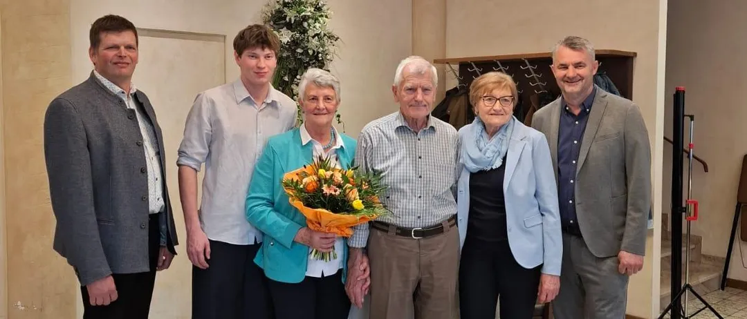 Eine Gruppe von Menschen steht zusammen in einem Raum. Eine ältere Frau hält einen Blumenstrauß. Der Mann und die Frau in der Mitte lächeln. Eine Kronleuchter hängt von der Decke. Eine Pflanze steht auf der linken Seite.