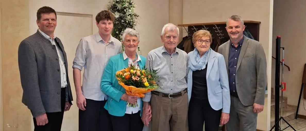Eine Gruppe von Menschen steht zusammen in einem Raum. Eine ältere Frau hält einen Blumenstrauß. Der Mann und die Frau in der Mitte lächeln. Eine Kronleuchter hängt von der Decke. Eine Pflanze steht auf der linken Seite.