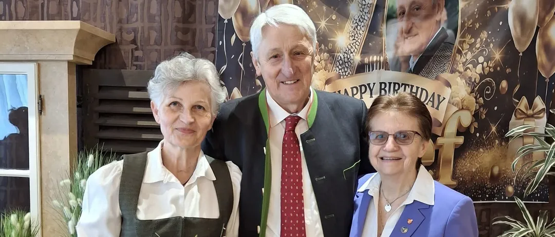 Drei Personen stehen vor einem Banner zur Feier eines 70. Geburtstags und lächeln. Der Mann trägt eine rote Krawatte und zwei Frauen tragen Kleider. Eine Frau hält einen Blumenstrauß. Ein Topf steht in der Nähe.