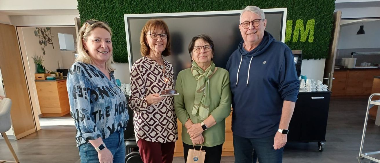 Vier Menschen stehen vor einer Wand mit einem großen Bildschirm. Eine Frau hält einen Teller mit einem Geburtstagskuchen. Eine andere Frau hält eine Tasche. Ein Mann hält ein Mikrofon.