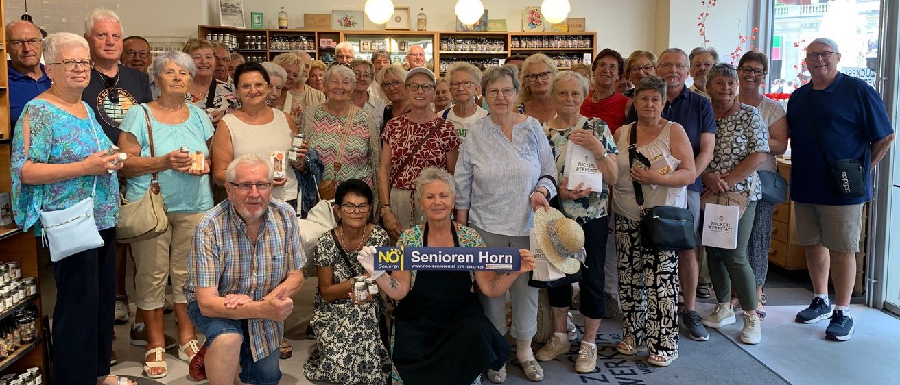 Eine Gruppe älterer Menschen versammelt sich in einem Geschäft, einige halten Gegenstände und eine Frau hält ein Schild mit der Aufschrift 'Senioren Horn'.