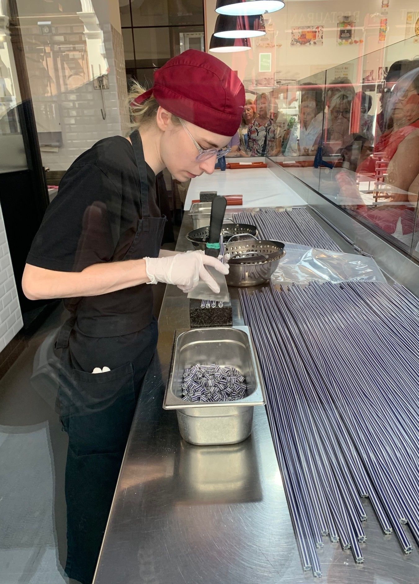 Eine Frau mit roter Haube und Brille arbeitet in einer Küche, hält ein Werkzeug und schaut auf ein Edelstahltablett mit kleinen Objekten.