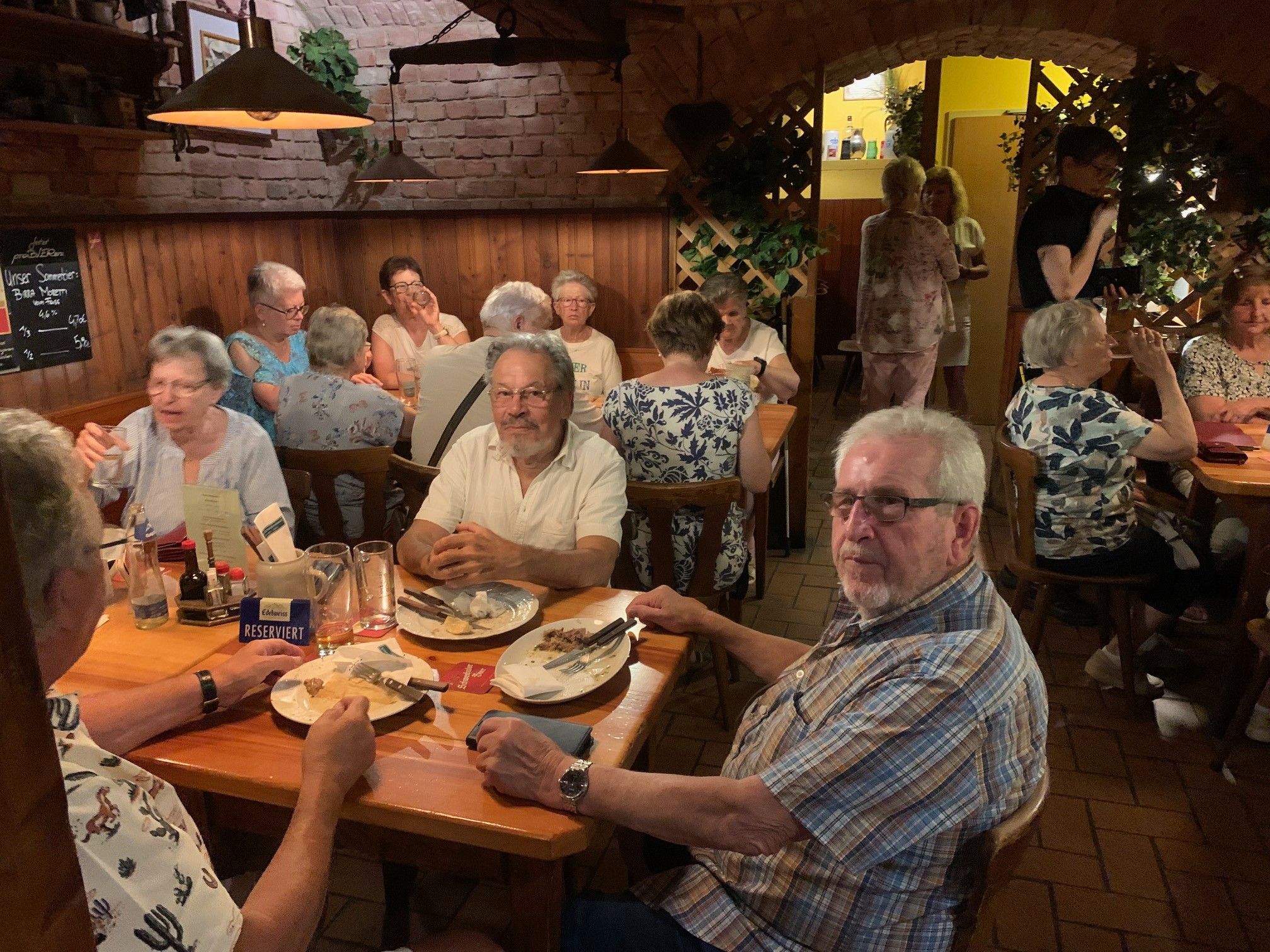 Eine Gruppe älterer Menschen sitzt an einem Restauranttisch, einige essen, während andere in Gespräche vertieft sind. Der Tisch hat Teller, Besteck und verschiedene Gewürze. Das Restaurant hat eine hölzerne Inneneinrichtung mit Ziegelwänden.