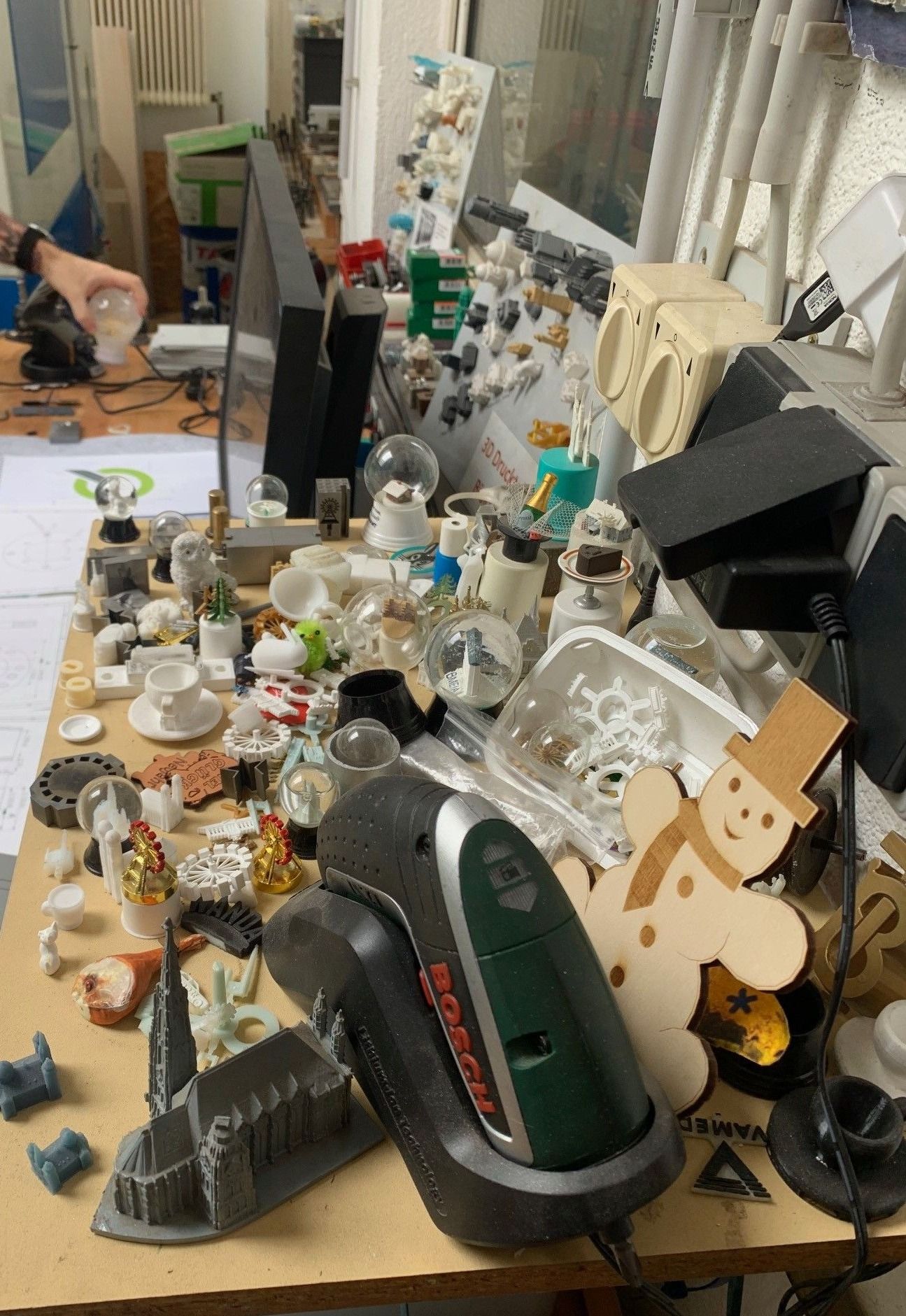 Ein überfüllter Schreibtisch mit verschiedenen 3D-gedruckten Objekten, einschließlich eines Schneemanns, einer Tasse und einer Flasche, zusammen mit einem Steckdosenverteiler und einer Bohrmaschine.