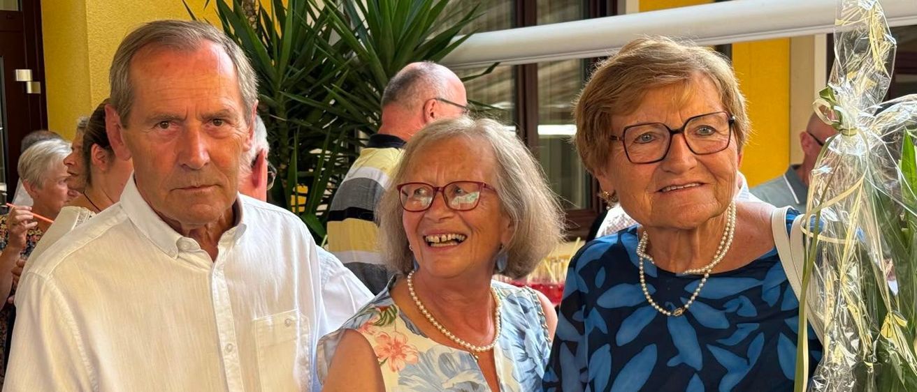 Drei Menschen stehen zusammen und lächeln für ein Foto. Der Mann links trägt ein weißes Hemd, die Frau in der Mitte trägt ein Blumenkleid und die Frau rechts trägt ein blaues Blumenoberteil. Sie sind draußen in der Nähe einer gelben Wand.