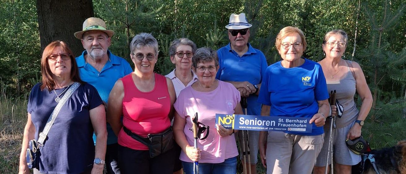 Eine Gruppe älterer Menschen, alle in Freizeitkleidung, stehen auf einem Pfad im Wald. Sie halten Wanderstöcke und lächeln für ein Foto. Ein Hund steht rechts.