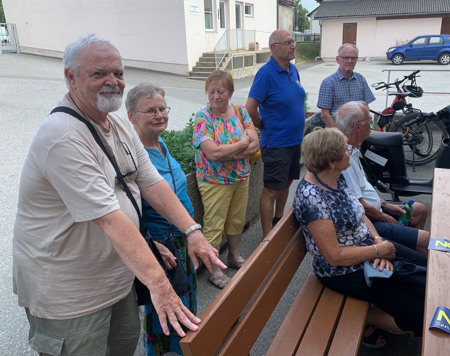 Eine Gruppe älterer Menschen, einige stehend und einige auf einer Bank sitzend, versammelt sich vor einem Gebäude mit einer Treppe und einem geparkten Auto.