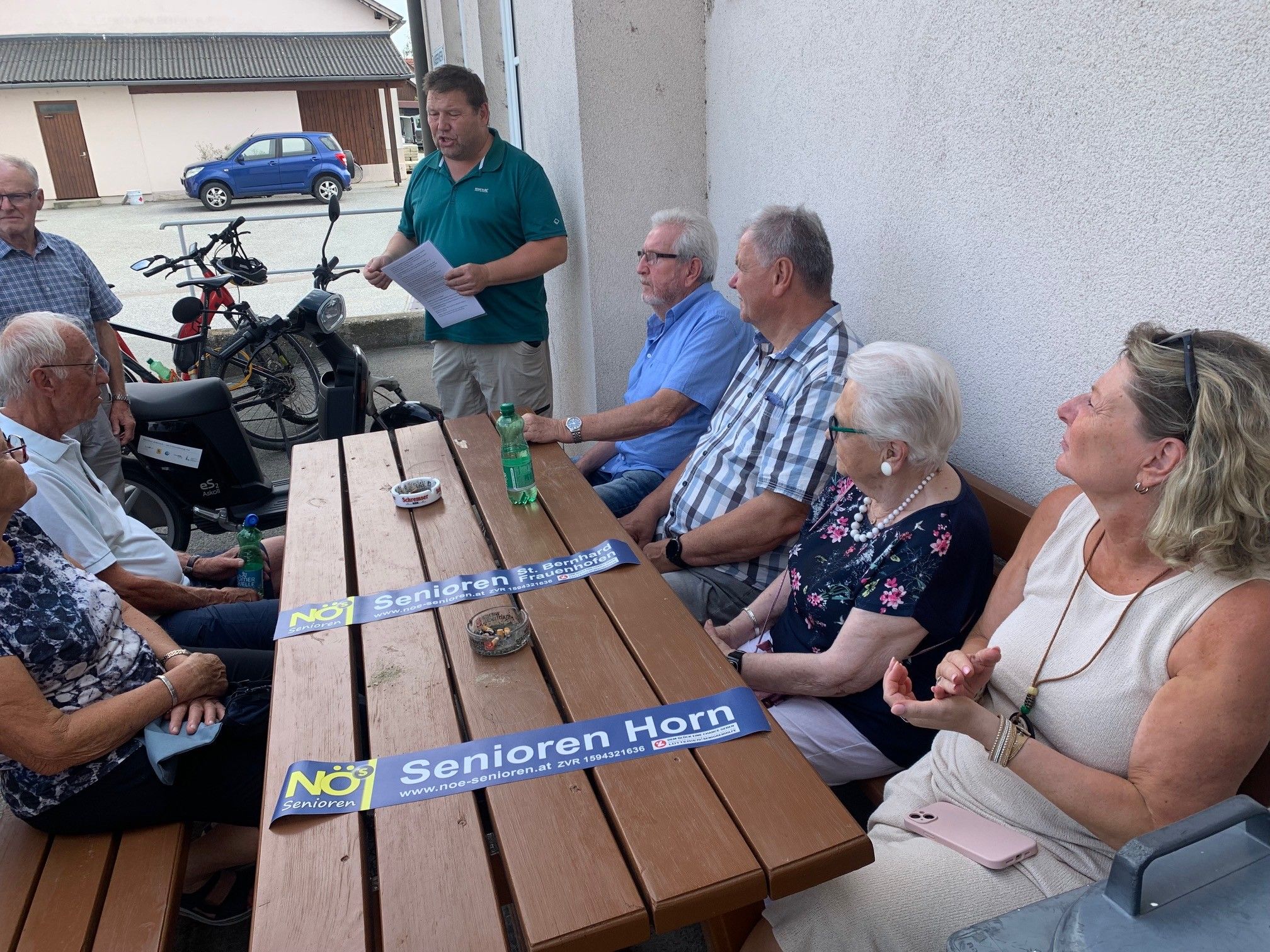 Eine Gruppe von Menschen sitzt an einem Holztisch, während ein Mann steht und spricht. Hinter ihnen sind Fahrräder geparkt. Banner mit dem Text 'Senioren Horn' liegen auf dem Tisch.