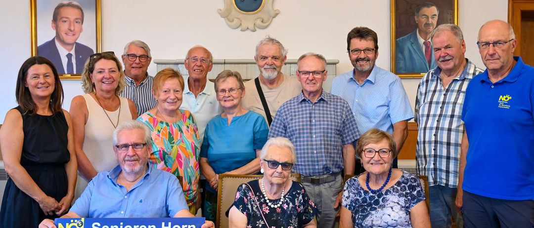 Eine Gruppe älterer Menschen posiert für ein Foto in einem Raum mit einem Holztisch, einem blauen Schild und einem Wappen an der Wand.