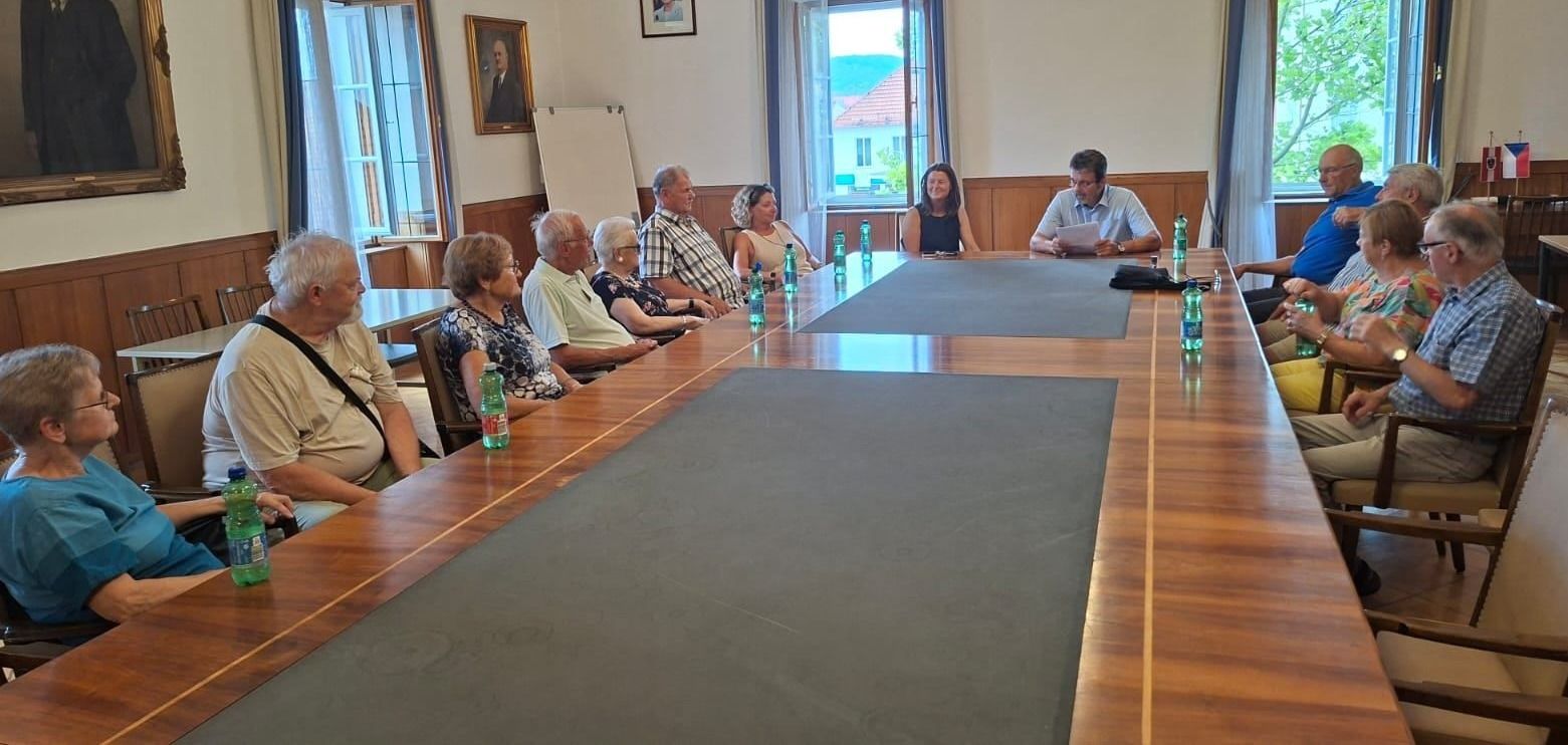 Eine Gruppe von Menschen sitzt um einen langen Tisch in einem Besprechungsraum, mit Flaschen und einer Tafel im Hintergrund.