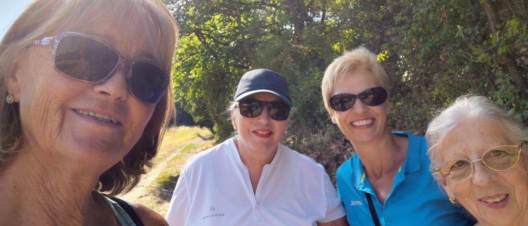 Vier Frauen lächeln und posieren für ein Foto im Wald. Die Frau auf der linken Seite trägt eine Sonnenbrille und einen grünen Riemen. Die Frau in der Mitte trägt ein weißes Hemd und eine blaue Kappe. Die Frau auf der rechten Seite trägt ein blaues Hemd. Die Frau ganz rechts trägt eine Brille.