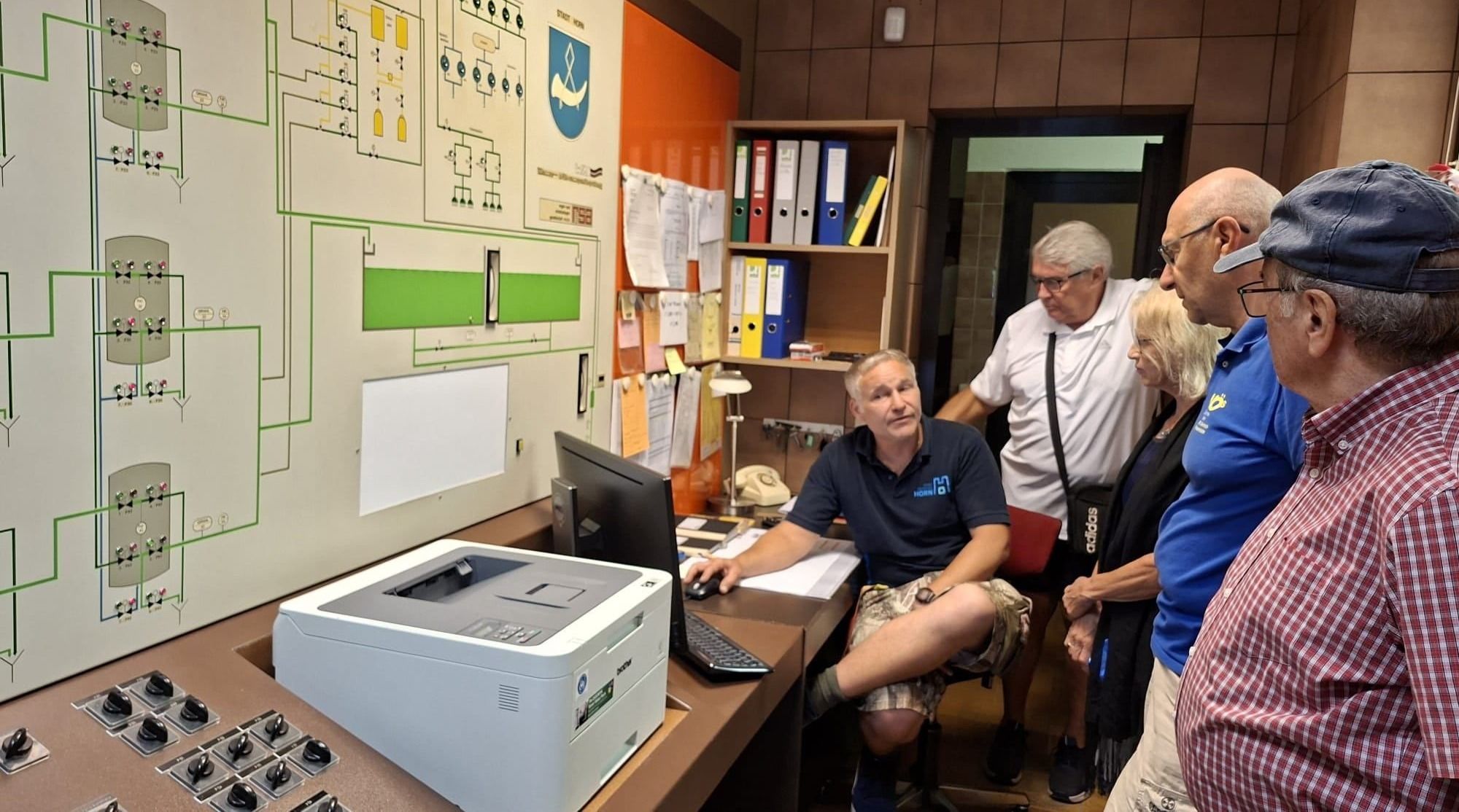 Drei Männer in einem Büro, einer sitzt an einem Schreibtisch mit einem Computer, ein anderer steht mit einer Tasche und ein dritter steht hinter dem sitzenden Mann.