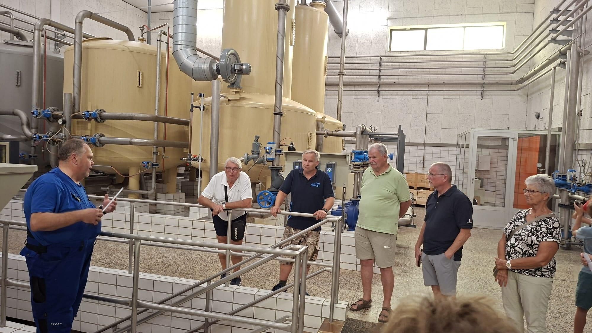 Vier Männer in Freizeitkleidung stehen in einer industriellen Umgebung, möglicherweise in einer Fabrik, mit großer Maschinen und Rohrleitungen im Hintergrund.