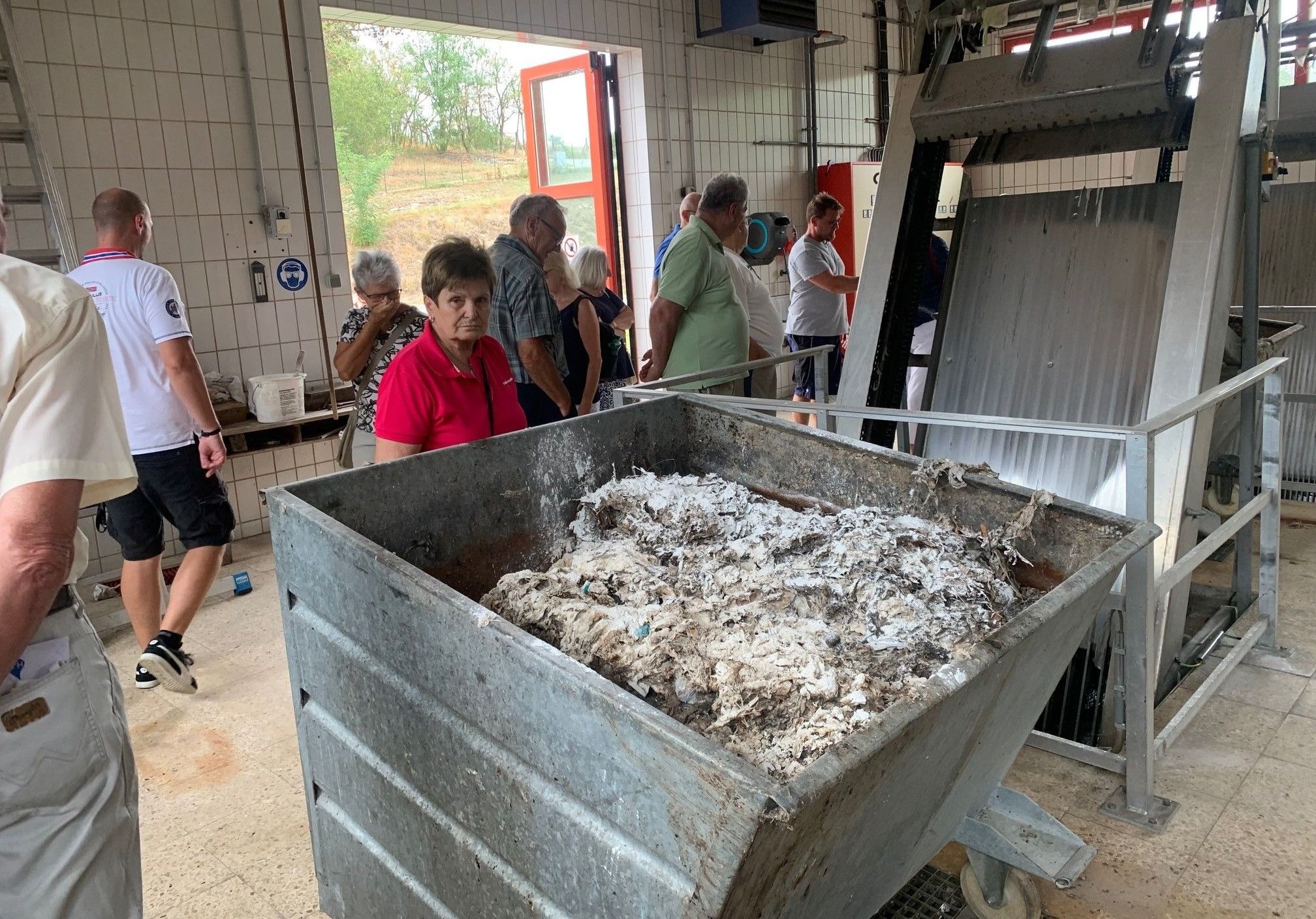 Eine Gruppe von Menschen steht in einem Raum mit einem großen Metallbehälter, der mit Wolle gefüllt ist. Der Raum hat geflieste Wände und eine offene Tür, die nach draußen führt.