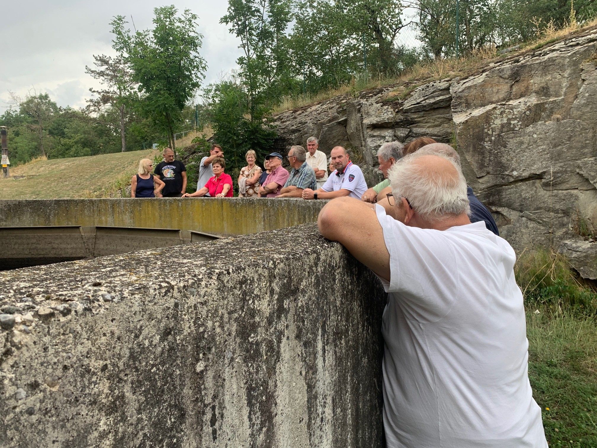 Eine Gruppe von Menschen, einige mit Brille, steht auf einer Betonmauer und beobachtet möglicherweise etwas. Die Mauer ist von einem felsigen Hügel mit Bäumen und Gras umgeben.