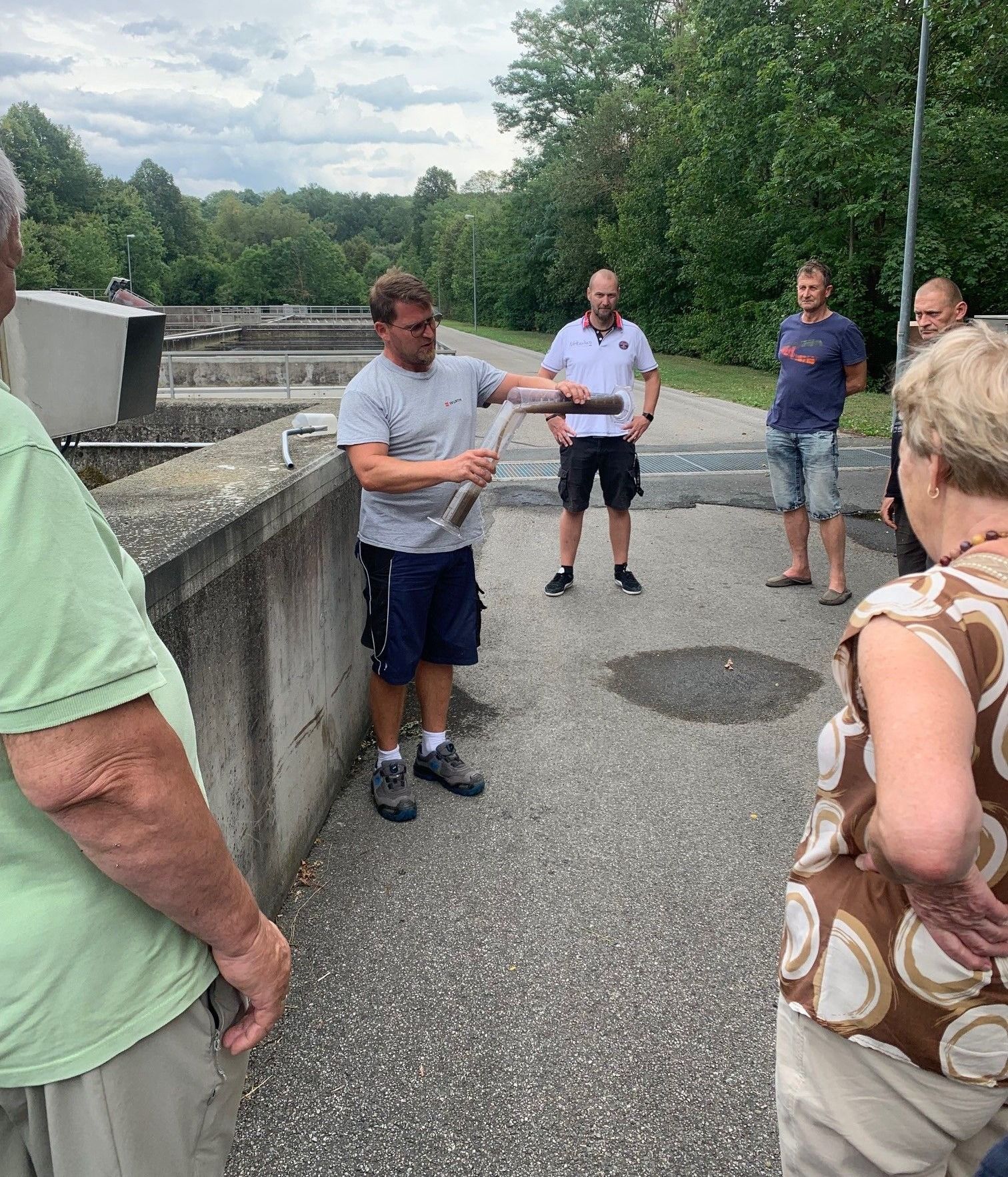Eine Gruppe von Menschen steht auf einer Betonbrücke, einer von ihnen hält eine lange Metallröhre. Sie alle schauen auf etwas vor sich. Dahinter befindet sich eine große Betonstruktur mit einem Gewässer.