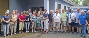 Eine Gruppe von Menschen, möglicherweise Senioren, steht vor einem Gebäude und posiert lächelnd für ein Foto. Sie tragen Freizeitkleidung und einige halten Papiere in der Hand.