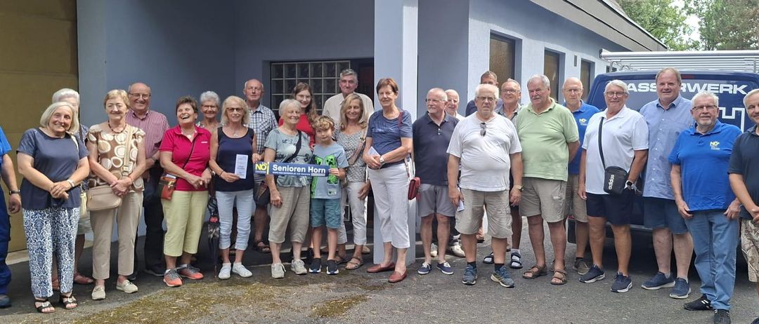 Eine Gruppe von Menschen, möglicherweise Senioren, steht vor einem Gebäude und posiert lächelnd für ein Foto. Sie tragen Freizeitkleidung und einige halten Papiere in der Hand.