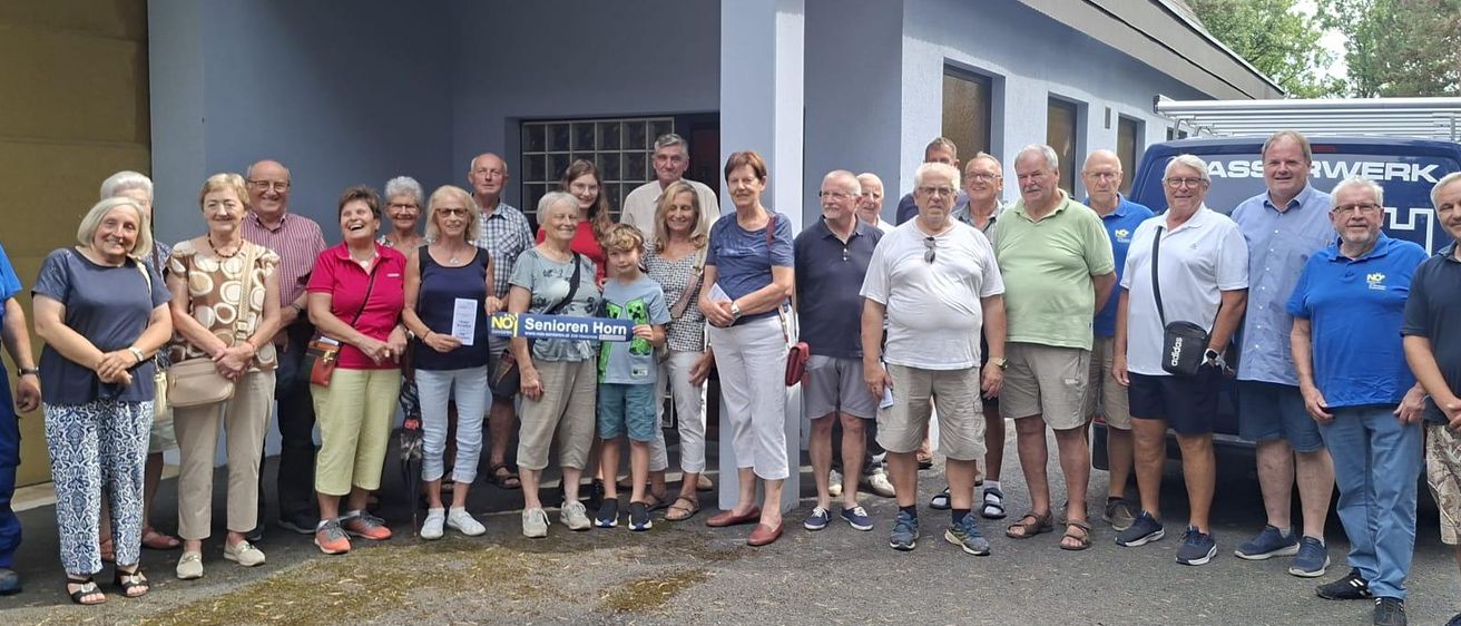 Eine Gruppe von Menschen, möglicherweise Senioren, steht vor einem Gebäude und posiert lächelnd für ein Foto. Sie tragen Freizeitkleidung und einige halten Papiere in der Hand.