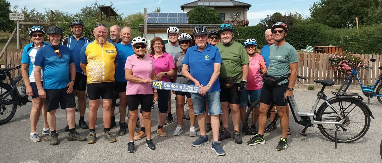 Eine Gruppe von Radfahrern in verschiedenfarbigen Trikots und Helmen posiert für ein Foto vor einem Haus mit Solarmodulen.