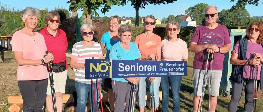 Eine Gruppe älterer Frauen und Männer steht zusammen auf einem Weg, hält Wanderstöcke in der Hand, mit einem blauen Schild, auf dem 'Senioren' steht. Sie lächeln und scheinen für ein Foto zu posieren.