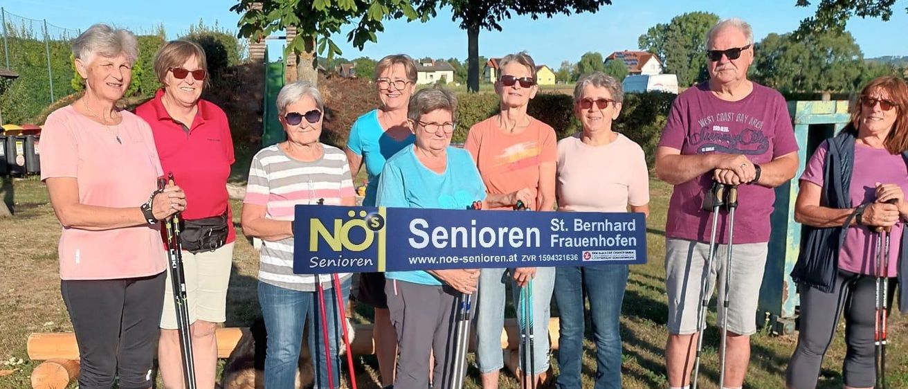 Eine Gruppe älterer Frauen und Männer steht zusammen auf einem Weg, hält Wanderstöcke in der Hand, mit einem blauen Schild, auf dem 'Senioren' steht. Sie lächeln und scheinen für ein Foto zu posieren.