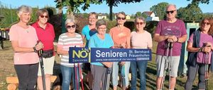 Eine Gruppe älterer Frauen und Männer steht zusammen auf einem Weg, hält Wanderstöcke in der Hand, mit einem blauen Schild, auf dem 'Senioren' steht. Sie lächeln und scheinen für ein Foto zu posieren.