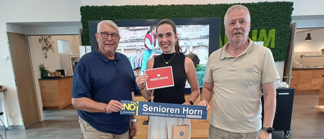Drei Personen stehen lächelnd zusammen, einer hält ein blaues Schild mit der Aufschrift 'Senioren Horn' und ein anderer ein rotes Schild mit dem Text 'Moser Reisen'. Sie befinden sich in einem Raum mit einer grünen Wand und einem großen Bildschirm dahinter.