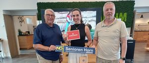 Drei Personen stehen lächelnd zusammen, einer hält ein blaues Schild mit der Aufschrift 'Senioren Horn' und ein anderer ein rotes Schild mit dem Text 'Moser Reisen'. Sie befinden sich in einem Raum mit einer grünen Wand und einem großen Bildschirm dahinter.