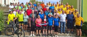 Eine Gruppe älterer Erwachsener, die in Fahrradausrüstung gekleidet sind, stehen auf Stufen mit einem grünen Gebäude im Hintergrund. Sie halten ein blaues Banner mit der Aufschrift 'Senioren Horn'.