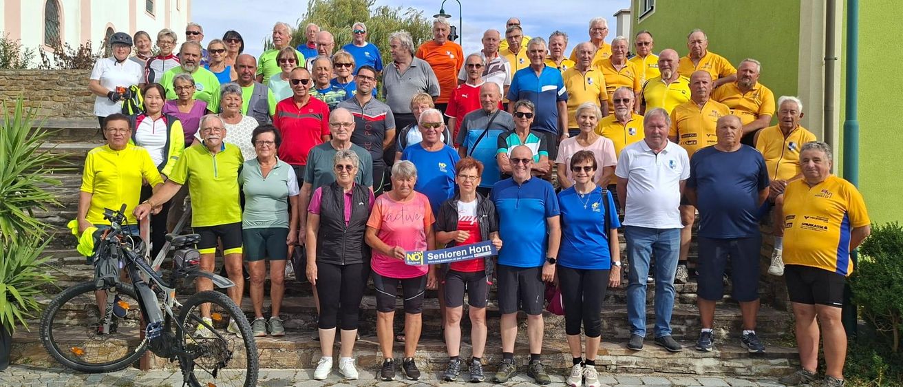 Eine Gruppe älterer Erwachsener, die in Fahrradausrüstung gekleidet sind, stehen auf Stufen mit einem grünen Gebäude im Hintergrund. Sie halten ein blaues Banner mit der Aufschrift 'Senioren Horn'.