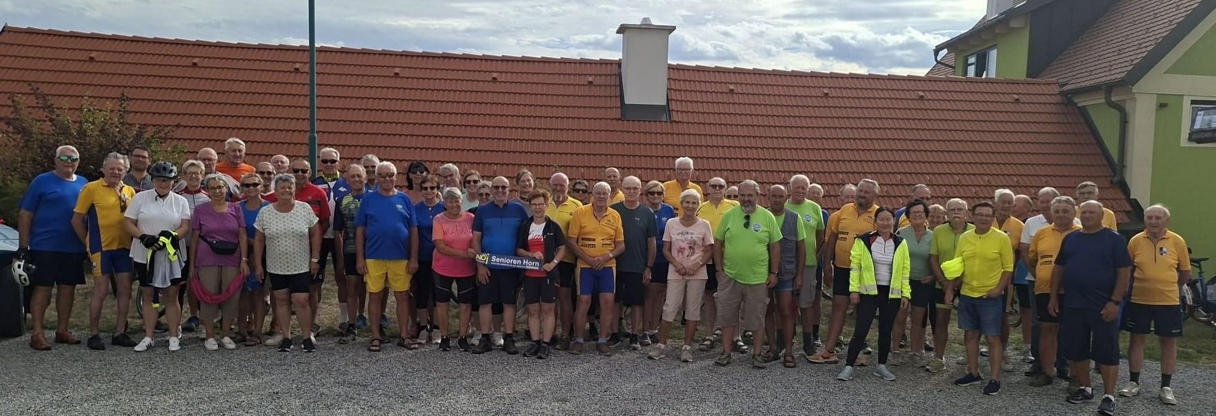 Eine Gruppe von Menschen in Fahrradbekleidung, die vor einem rotgedeckten Haus stehen, einige mit Sonnenbrillen und einem Banner.