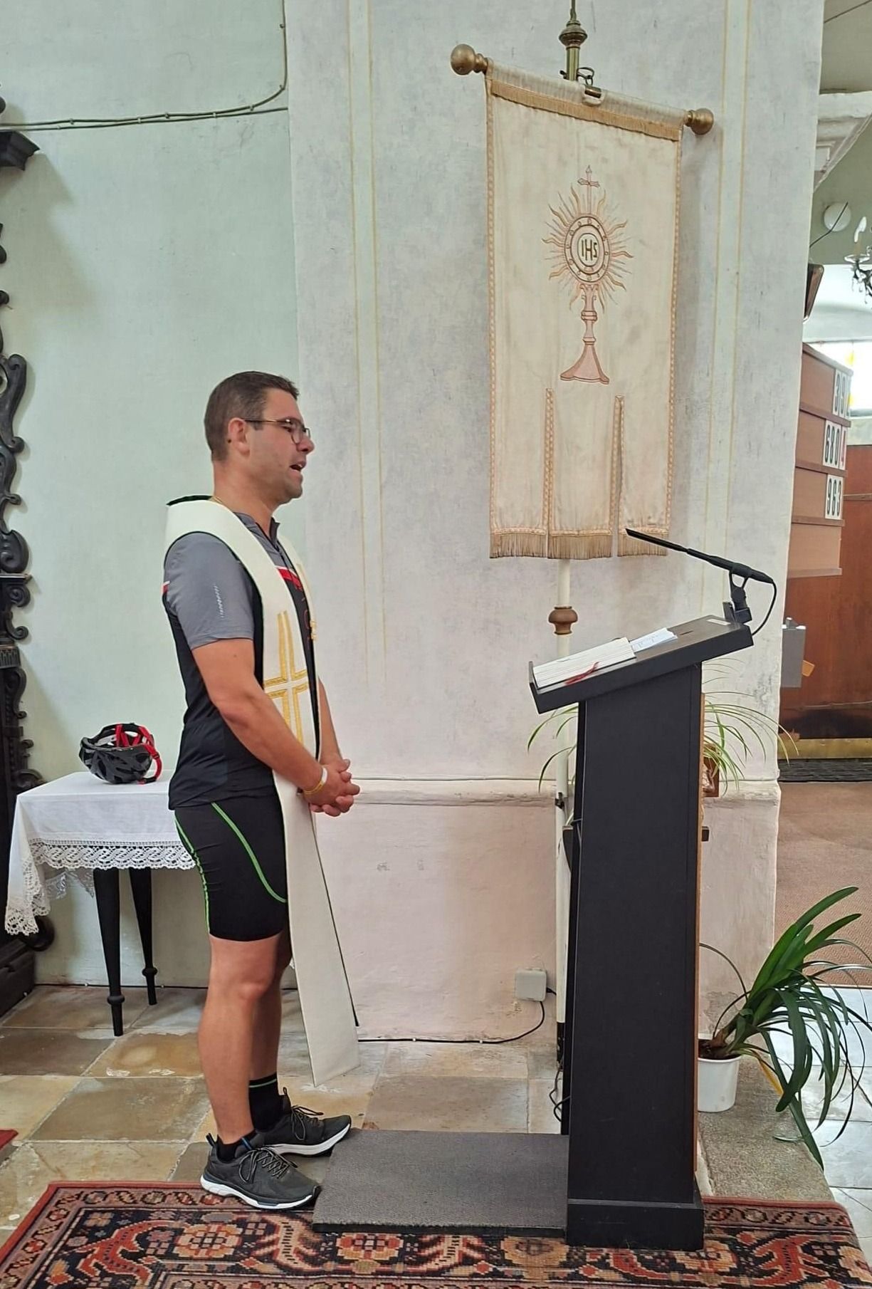 Ein Mann mit einer weißen Stola steht vor einem Podest in einer Kirche, mit einem Mikrofon und einem Banner hinter ihm.
