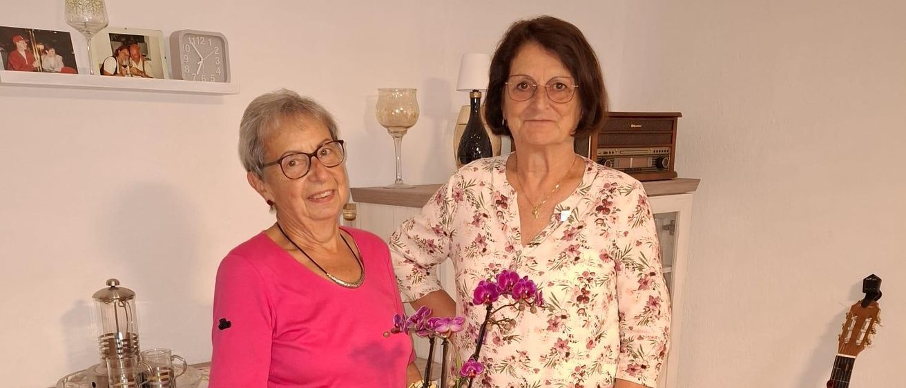 Zwei Frauen stehen in einem Raum, eine hält eine Topfpflanze und lächelt. Dahinter steht ein Schrank mit einer Lampe und einer Gitarre.