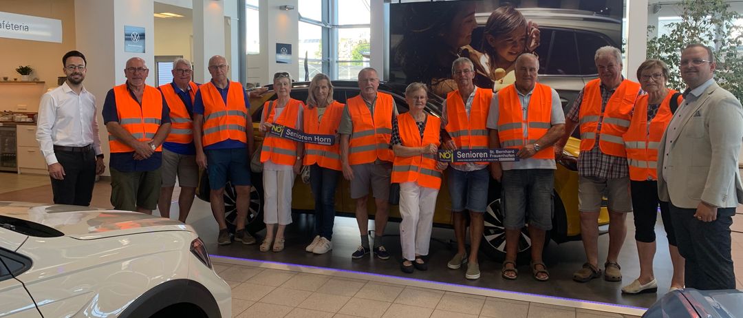 Eine Gruppe älterer Personen, die orange Westen tragen, posiert für ein Foto in einer Autoausstellung. Sie stehen vor einem gelben Auto mit einem großen Poster im Hintergrund.