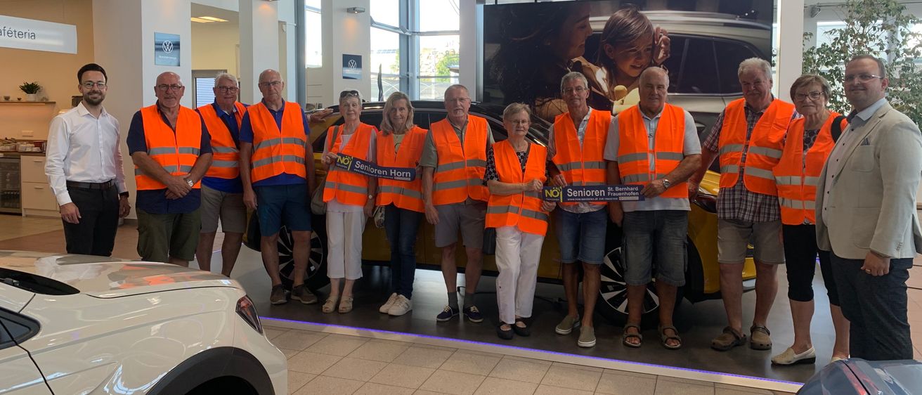 Eine Gruppe älterer Personen, die orange Westen tragen, posiert für ein Foto in einer Autoausstellung. Sie stehen vor einem gelben Auto mit einem großen Poster im Hintergrund.