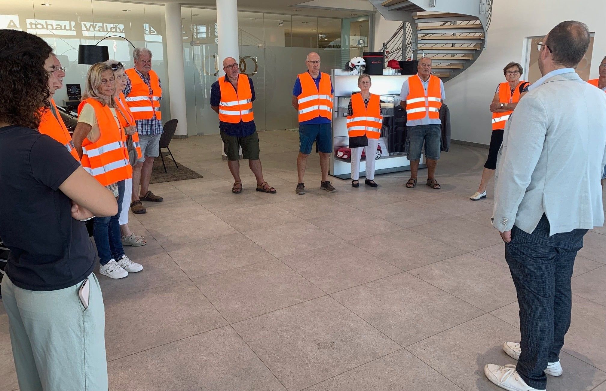Eine Gruppe von Menschen in orangefarbenen Sicherheitswesten steht in einem modernen Gebäude mit gefliesten Böden und einer Treppe im Hintergrund.