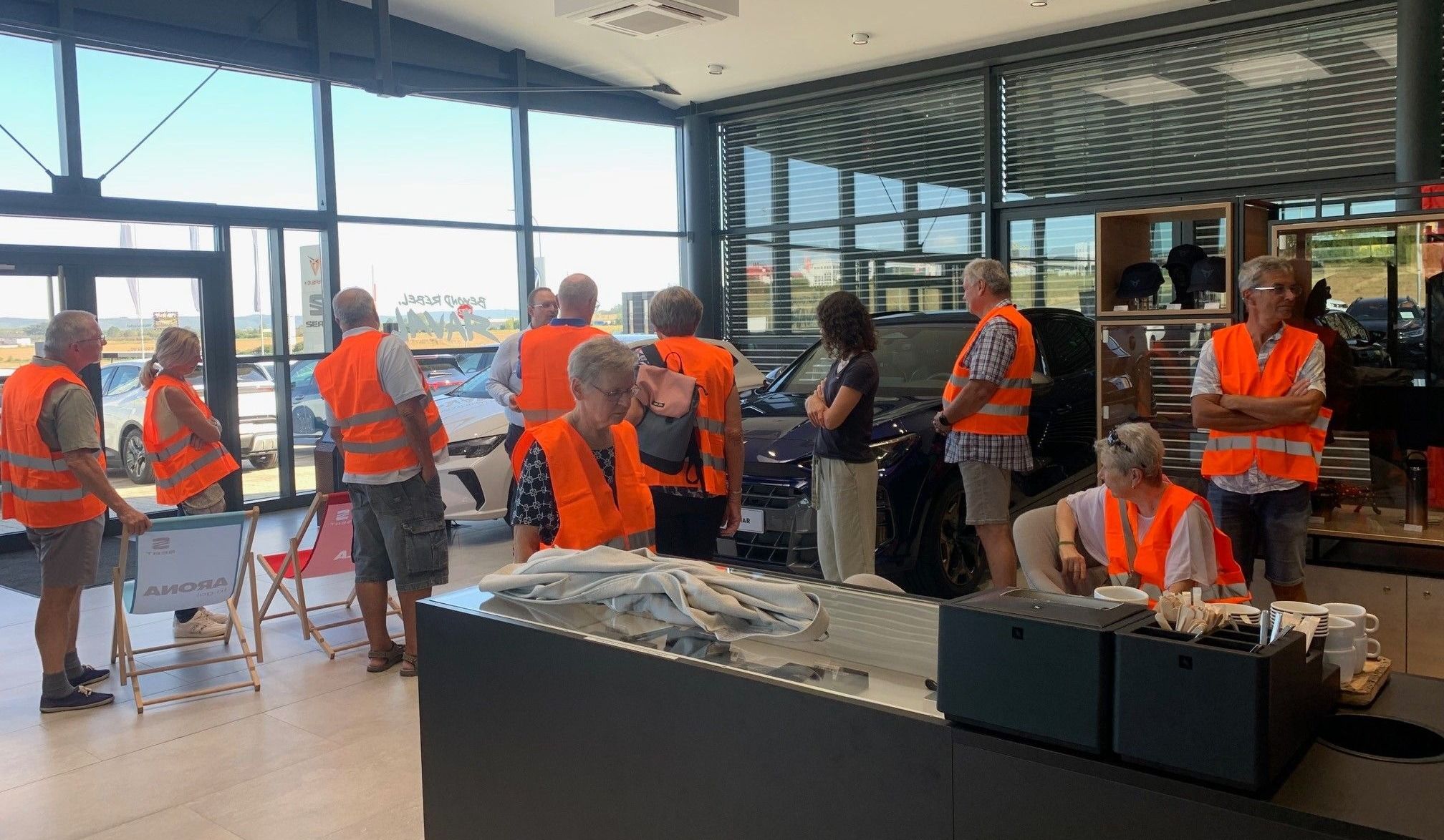 Eine Gruppe von Menschen in orangenen Westen steht in einem Autohaus, mit einem schwarzen Auto im Vordergrund und einem weißen Auto im Hintergrund.