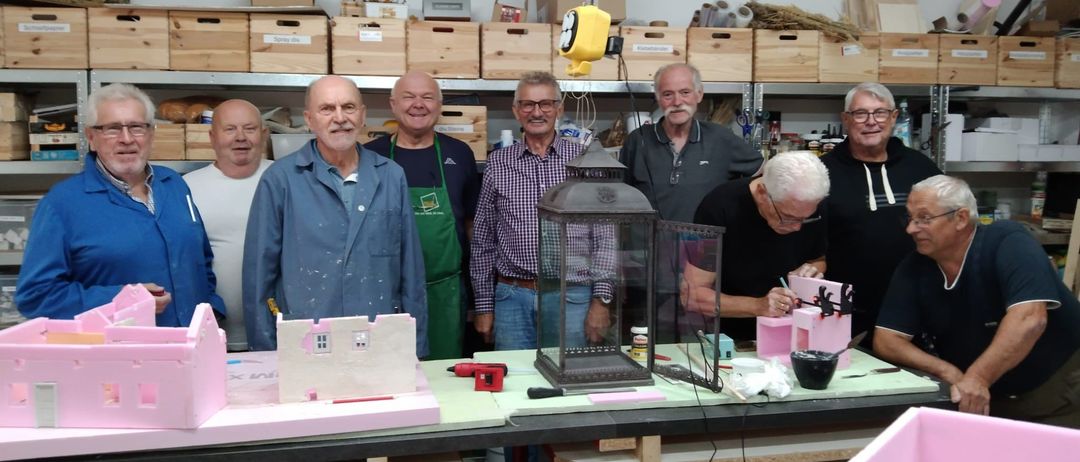 Eine Gruppe älterer Männer in einer Werkstatt, die an einem Projekt arbeiten. Sie stehen um einen Tisch herum mit verschiedenen Werkzeugen und Materialien. Einige tragen Schürzen.