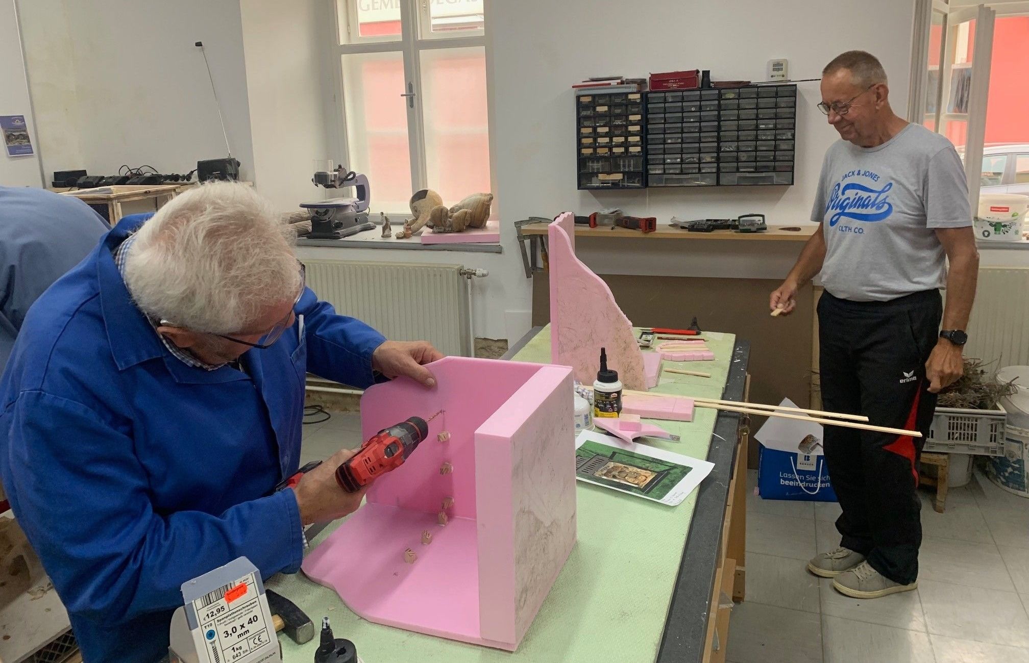 Ein älterer Mann mit Brille und blauer Jacke benutzt eine Bohrmaschine, um an einem rosa Objekt auf einem grünen Tisch zu arbeiten. Ein anderer Mann steht in der Nähe und hält ein Stück Holz.