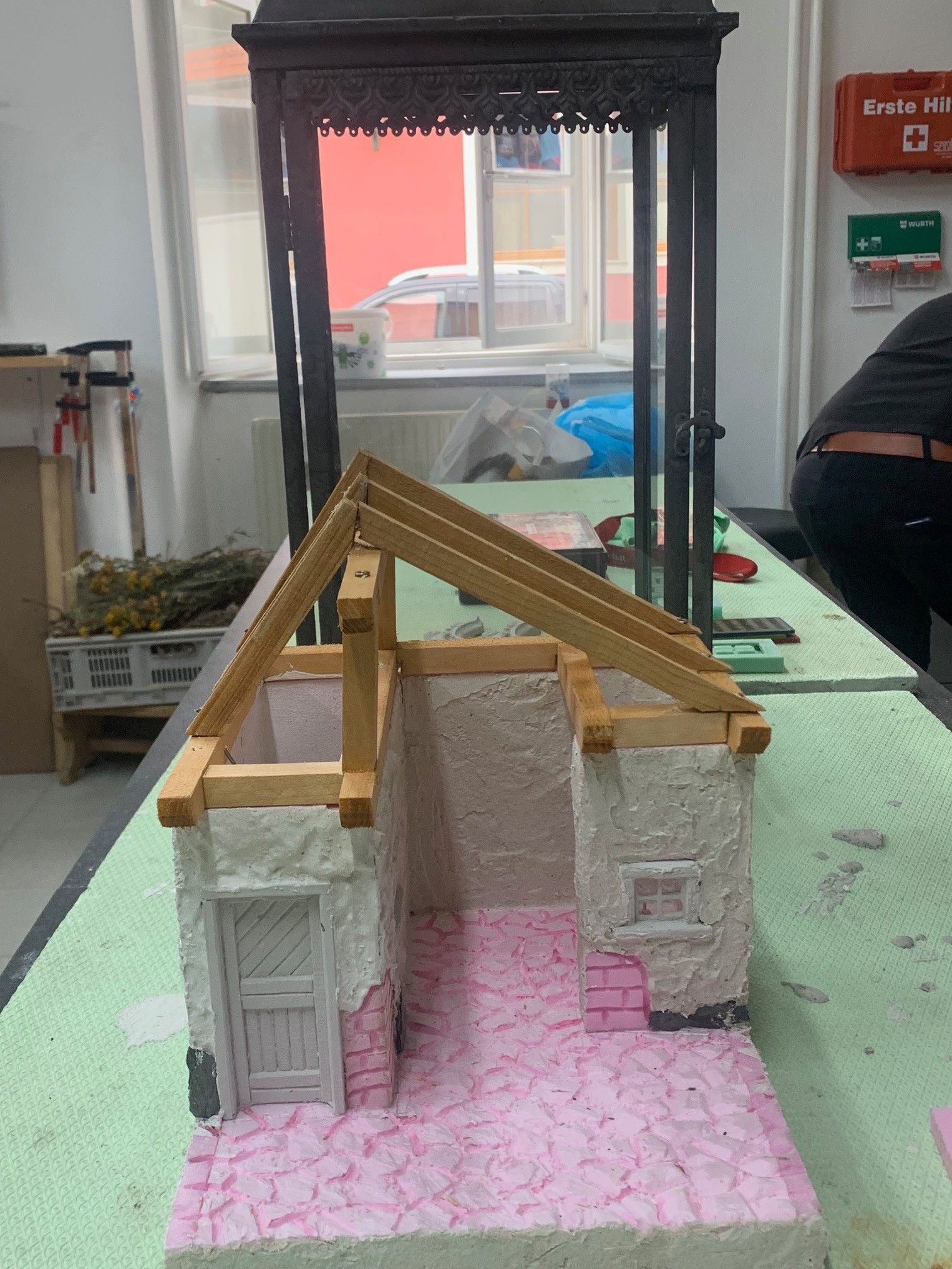 Ein kleines Modellhaus mit einem Holzdach und rosa Wänden wird auf einer grünen Oberfläche gebaut. Werkzeuge und Materialien sind verstreut.