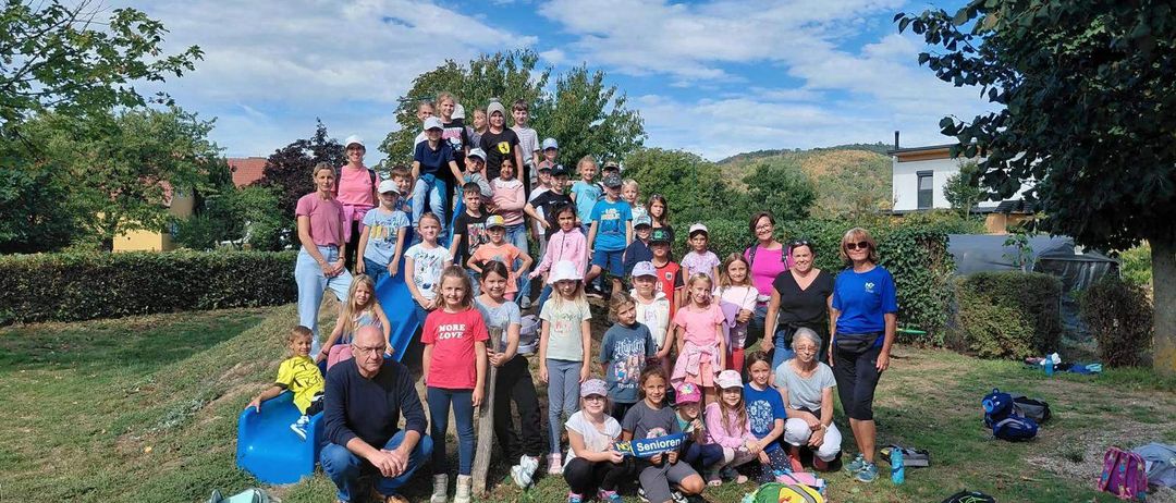 Eine Gruppe von Kindern und Erwachsenen posiert für ein Foto in einem Park mit einer blauen Rutsche und Bäumen im Hintergrund. Einige tragen Hüte und Turnschuhe, während andere ein Schild halten, das 'Senioren' besagt.