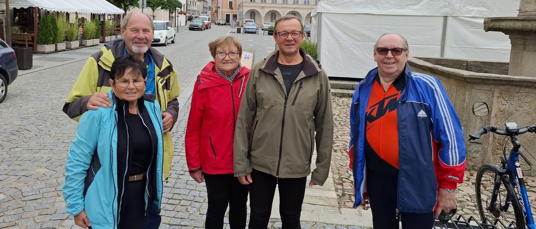 Fünf Personen stehen auf einer Kopfsteinpflasterstraße vor einem Gebäude mit einem Glockenturm. Sie lächeln und scheinen für ein Foto zu posieren.