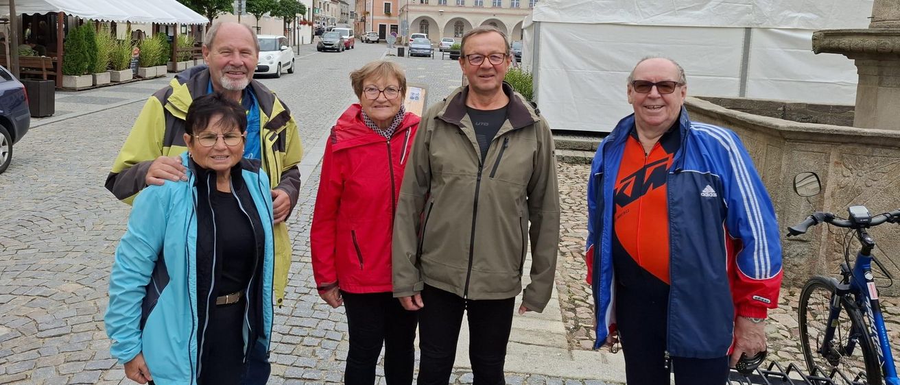 Fünf Personen stehen auf einer Kopfsteinpflasterstraße vor einem Gebäude mit einem Glockenturm. Sie lächeln und scheinen für ein Foto zu posieren.