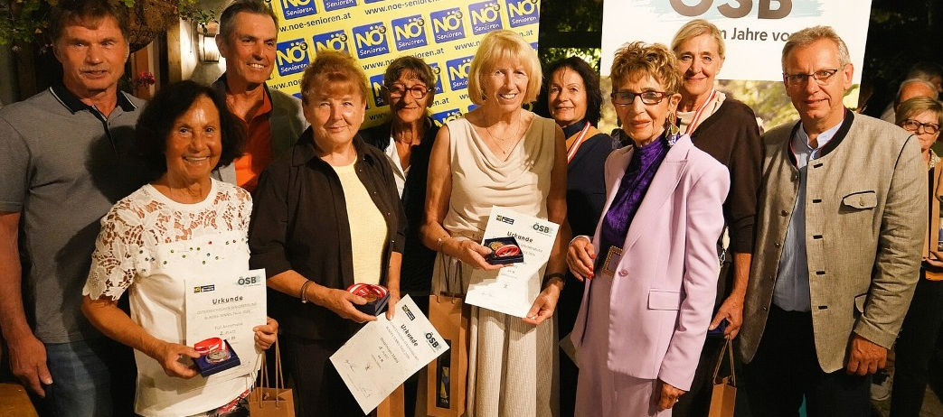 Eine Gruppe älterer Menschen steht lächelnd zusammen und hält Auszeichnungen. Sie sind in verschiedenen Outfits gekleidet und einige tragen Brillen. Sie stehen vor einem gelben Banner mit dem Text 'NEIN' und einem Logo.