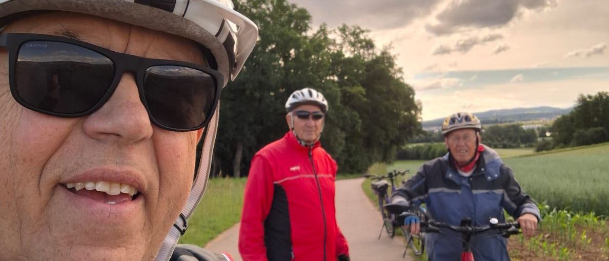 Drei Männer fahren auf einem Pfad Fahrrad. Ein Mann vorne trägt Helm und Sonnenbrille, ein anderer Mann im roten Mantel dahinter und ein dritter Mann mit Fahrrad.